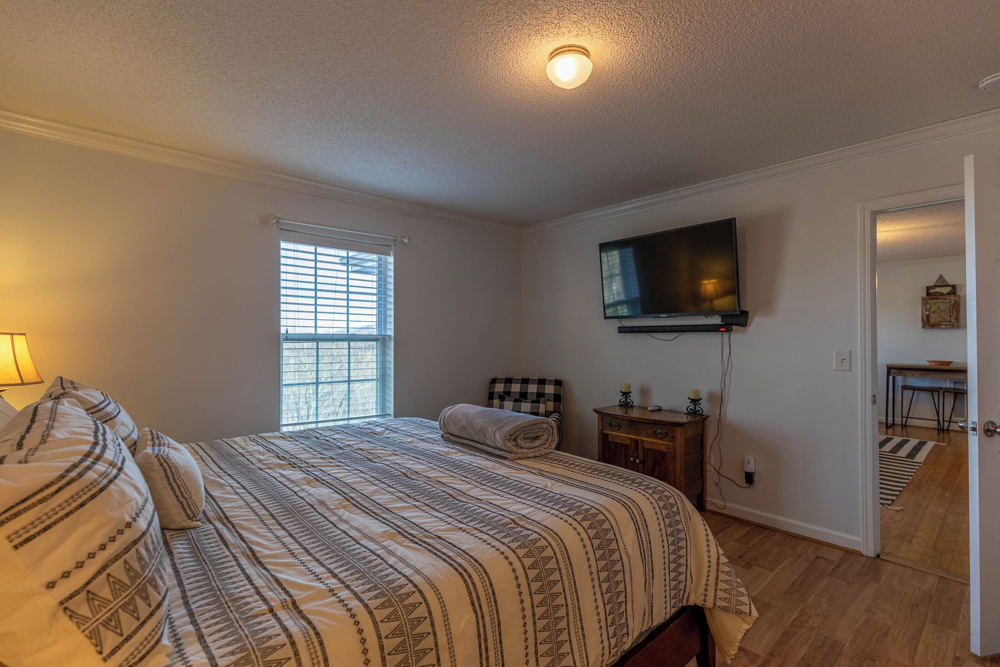A bedroom with a bed, a television mounted on the wall, a window with blinds, and a door leading to another room.
