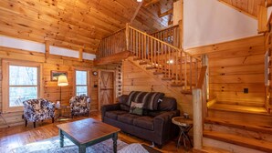 A wooden cabin interior with a staircase, a sofa, two armchairs, a coffee table, and a rug. - Whiskey Creek Cabin Peaceful Creekside Cabin With hot tub and Game Room (Sugar Grove)