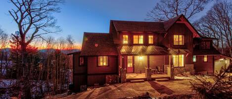 A large, multi-story wooden house with a garage, surrounded by trees and a snowy landscape.