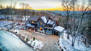 A large, dark-colored house with multiple windows and a garage, surrounded by a snowy landscape and trees.