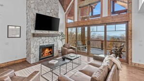 A spacious living room with a stone fireplace, leather furniture, and large windows offering a view of the outdoors.
