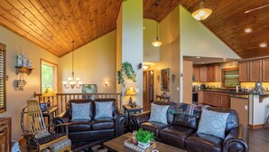 A spacious living room with a high ceiling, leather sofas, a wooden rocking chair, and a kitchen in the background. - Bearadise Cabin - Pet Friendly, Great Views, Firepit and Yard on Sugar Mountain, 2 Car Garage (Banner Elk)