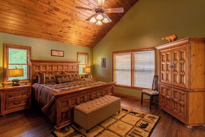 Une chambre avec un plafond en bois, un grand lit, une table de chevet en bois, un banc, une armoire et un ventilateur de plafond.