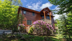 A wooden cabin with a balcony, surrounded by lush greenery and flowering bushes. - Bearadise Cabin - Pet Friendly, Great Views, Firepit and Yard on Sugar Mountain, 2 Car Garage (Banner Elk)