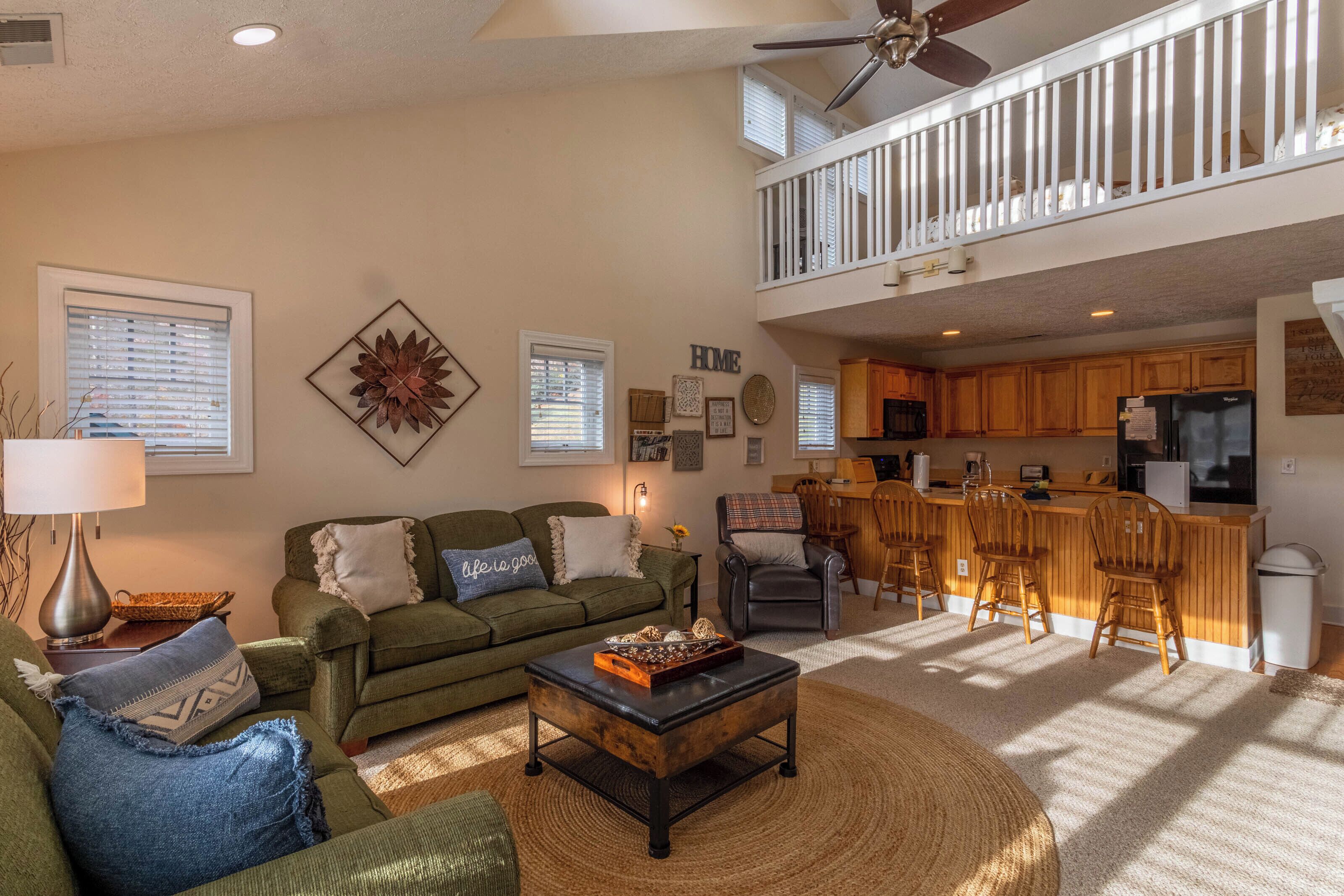 A spacious living room with a green sofa, a coffee table, and a kitchen area with wooden cabinets and a bar.