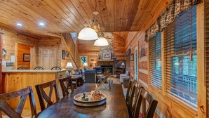 Interior de una cabaña de madera con comedor, chimenea y ventanas amplias.