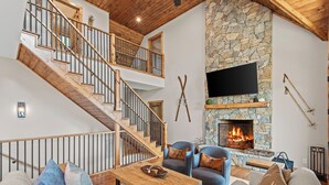 A modern living room with a stone fireplace, a flat-screen TV, and a wooden staircase. - Grandview Pointe (Banner Elk)