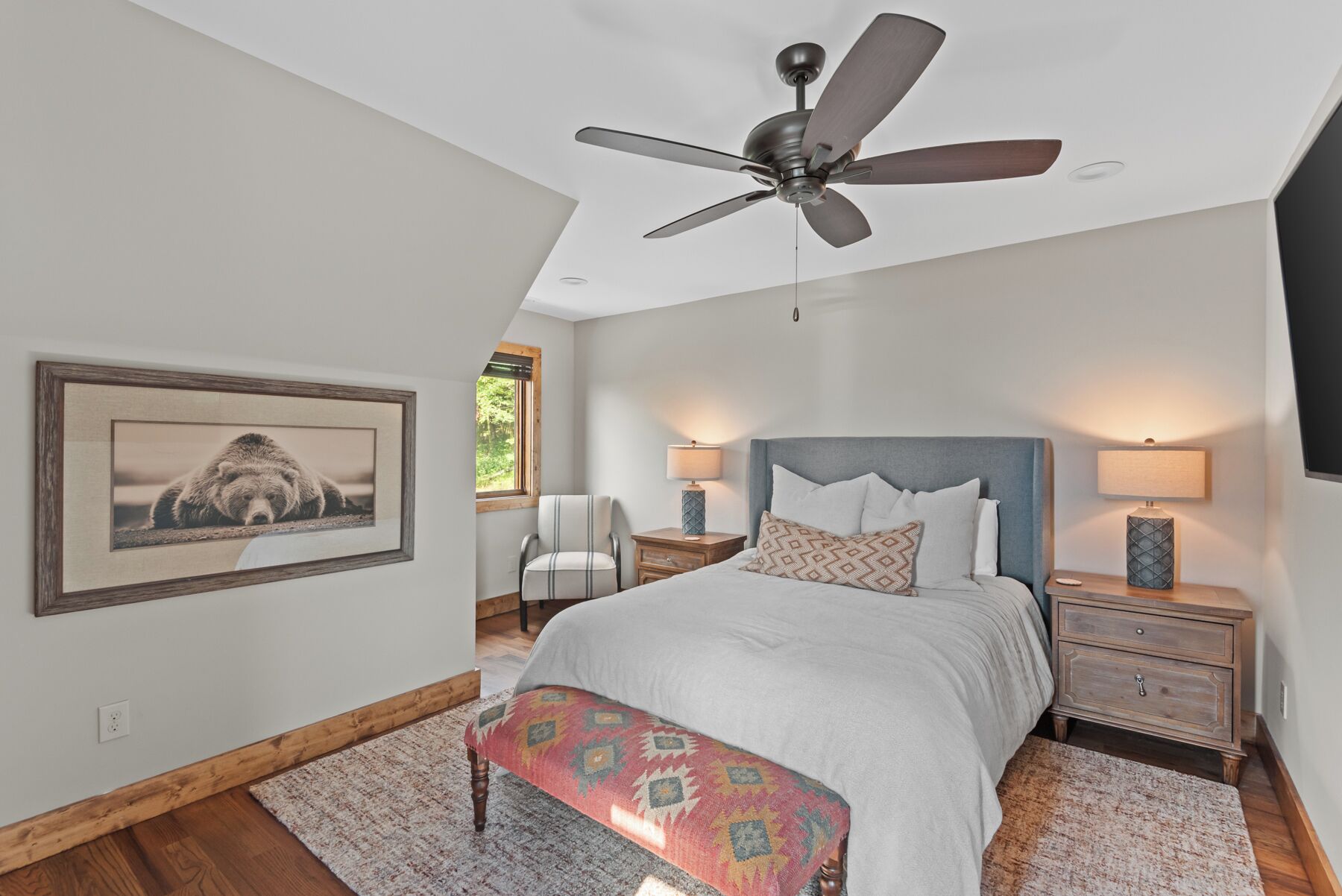 A bedroom with a bed, a ceiling fan, a nightstand, a chair, a lamp, a rug, and a framed picture on the wall.