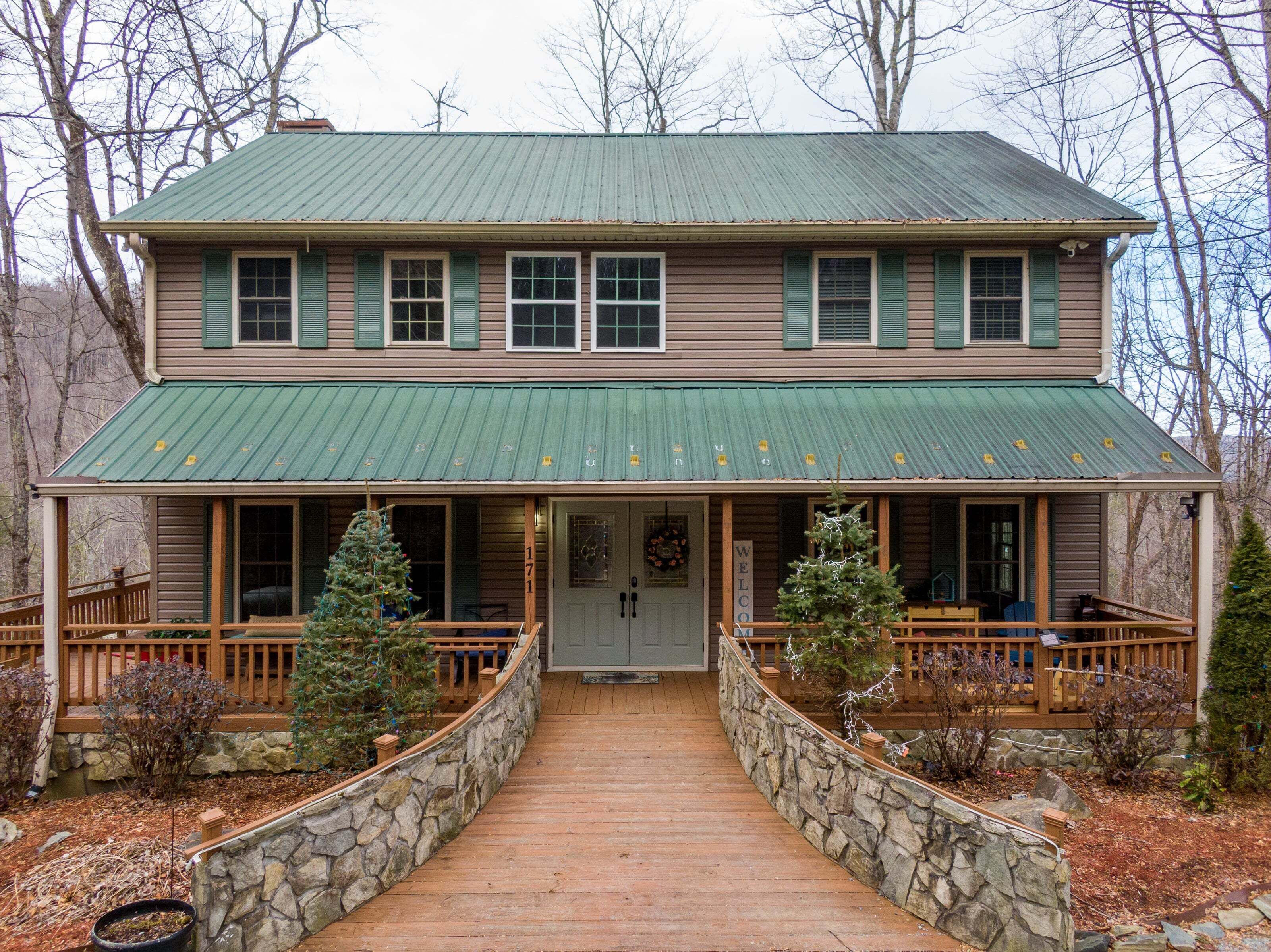 A two-story wooden house with a green metal roof, a stone pathway leading to the front door, and a welcoming sign.