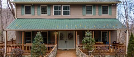 A two-story wooden house with a green metal roof, a stone pathway leading to the front door, and a welcoming sign.