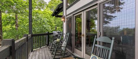 A wooden deck with rocking chairs and a grill, overlooking a forested area.