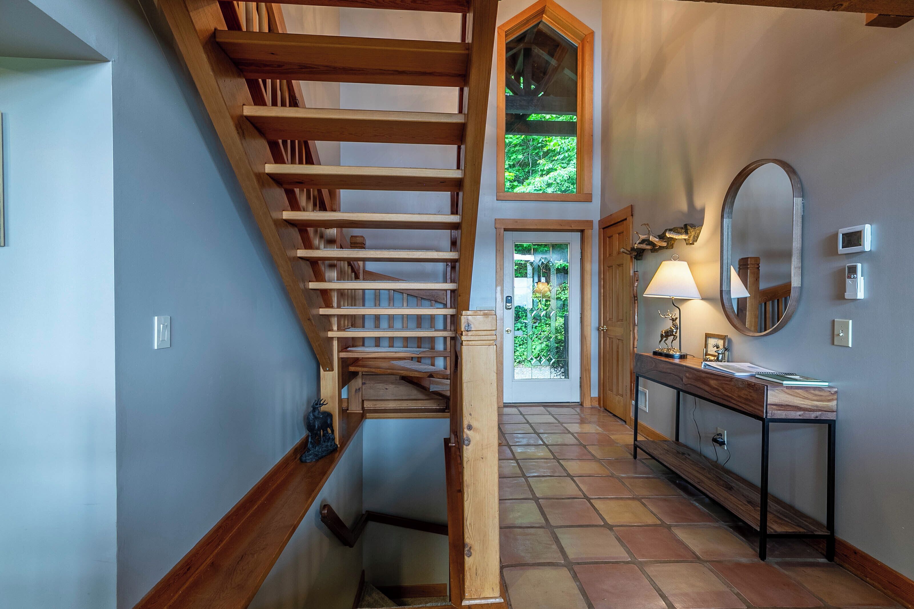 Eine Treppe führt zu einer zweiten Etage, ein Konsolentisch mit Spiegel und eine Tür, durch die man Grünflächen sieht.