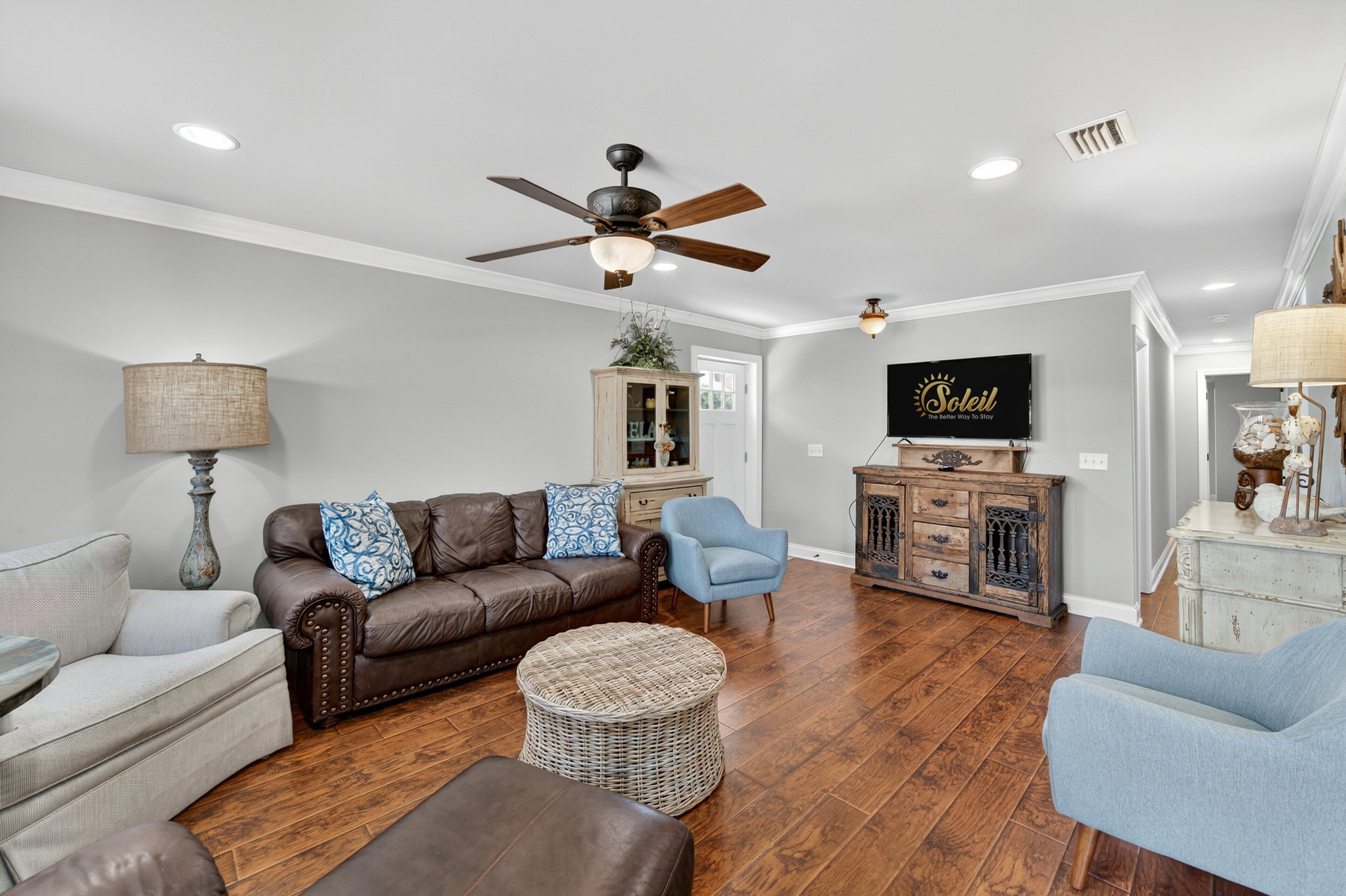 A spacious living room with a brown leather sofa, a blue armchair, and a wicker ottoman. A ceiling fan and a television are visible in the background.