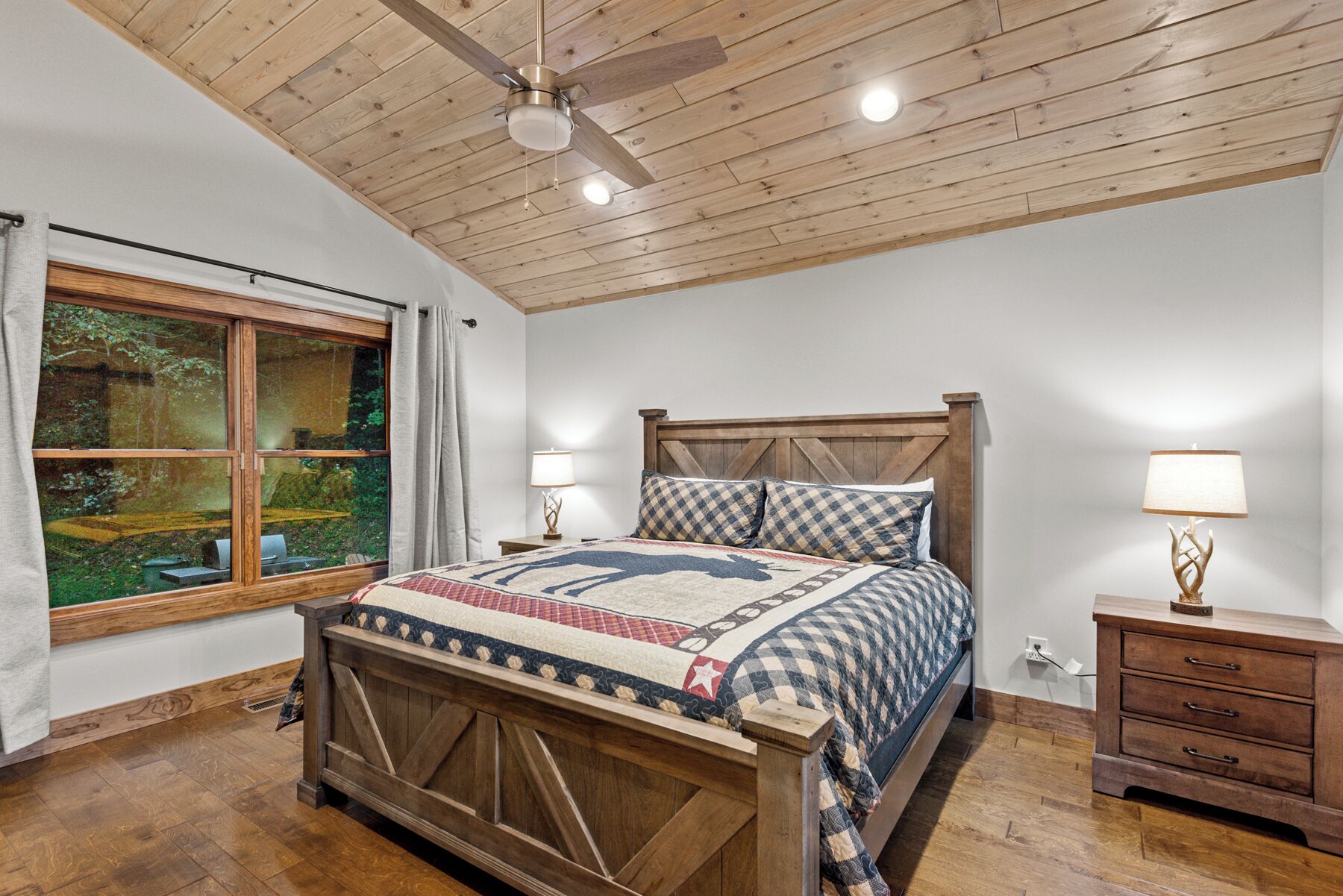 A bedroom with a wooden ceiling, a large window, a bed with a checkered quilt, bedside lamps, and a wooden nightstand.