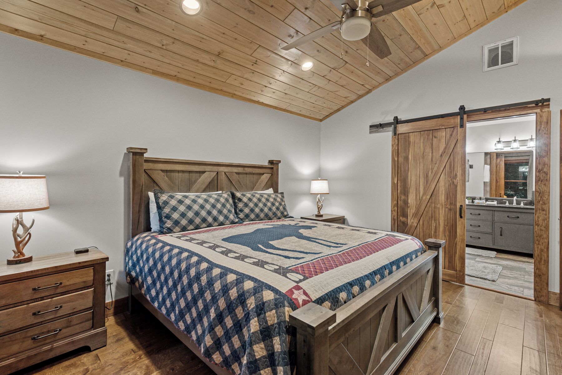 A bedroom with a wooden ceiling, a bed with a quilt, bedside lamps, a wooden nightstand, and a wardrobe.