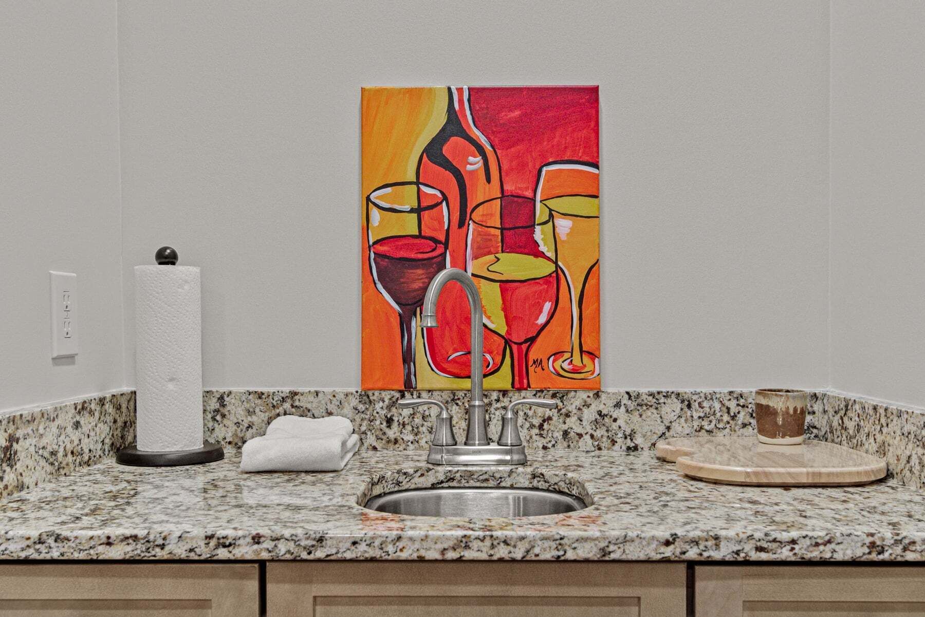Una cocina con encimera de granito, pileta de acero inoxidable, una toalla y un cuadro con copas de vino.