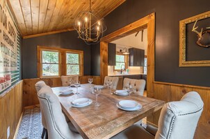 Un comedor con una mesa de madera dispuesta para seis personas, rodeada de sillas tapizadas. Un candelabro cuelga del techo y hay un adorno de cuernos enmarcado en la pared.