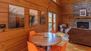 Een houten cabin interieur met een ronde houten tafel, oranje stoelen, een stenen openhaard en ingelijste kunstwerken aan de muren.