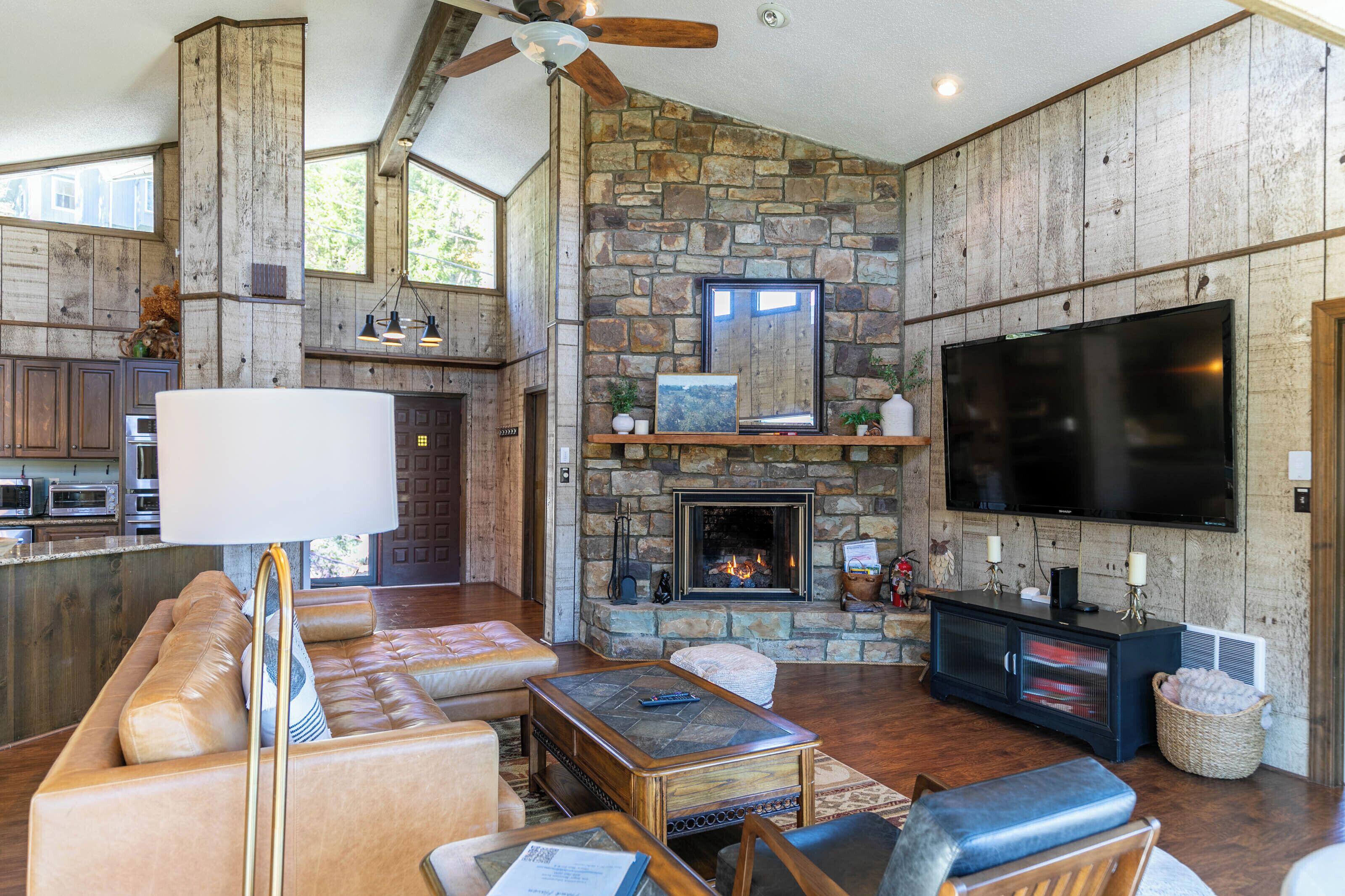 A spacious living area with a stone fireplace, a large flat-screen TV, leather seating, and a wooden ceiling fan.