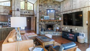 A spacious living area with a stone fireplace, a large flat-screen TV, leather seating, and a wooden ceiling fan. - Hawk Haven (Banner Elk)