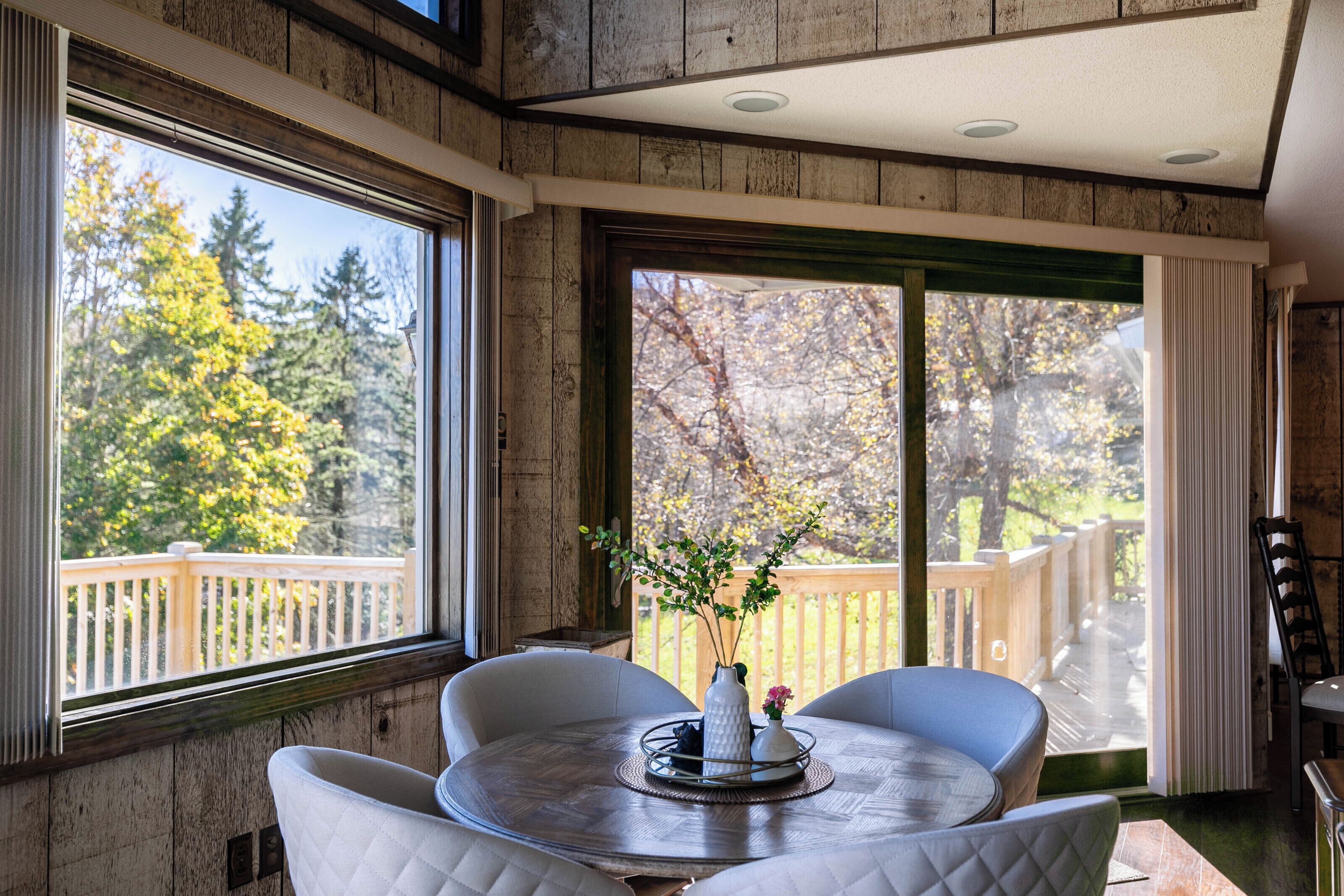 A room with a round table, a vase with flowers, and a view of a wooden deck and trees.