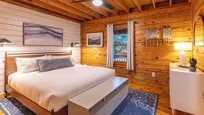 Un dormitorio con paneles de madera, una cama, un ventilador de techo, una ventana con cortinas, una mesita de noche con lámpara y un cuadro colgado en la pared.