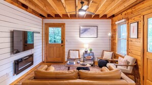 Interior acogedor de una cabaña con techo de madera, televisor, chimenea, sofás de cuero y un mapa enmarcado en la pared.