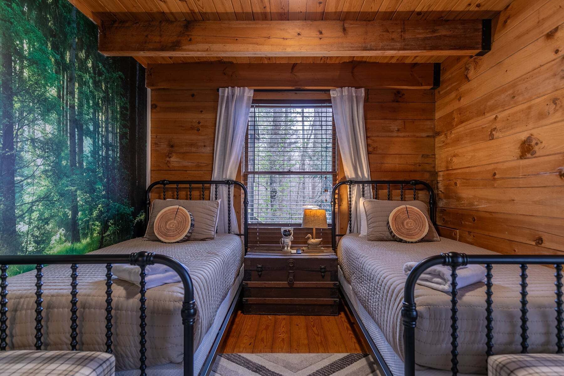 A cabin with two beds, a window with curtains, a small table, and a forest mural on the wall.