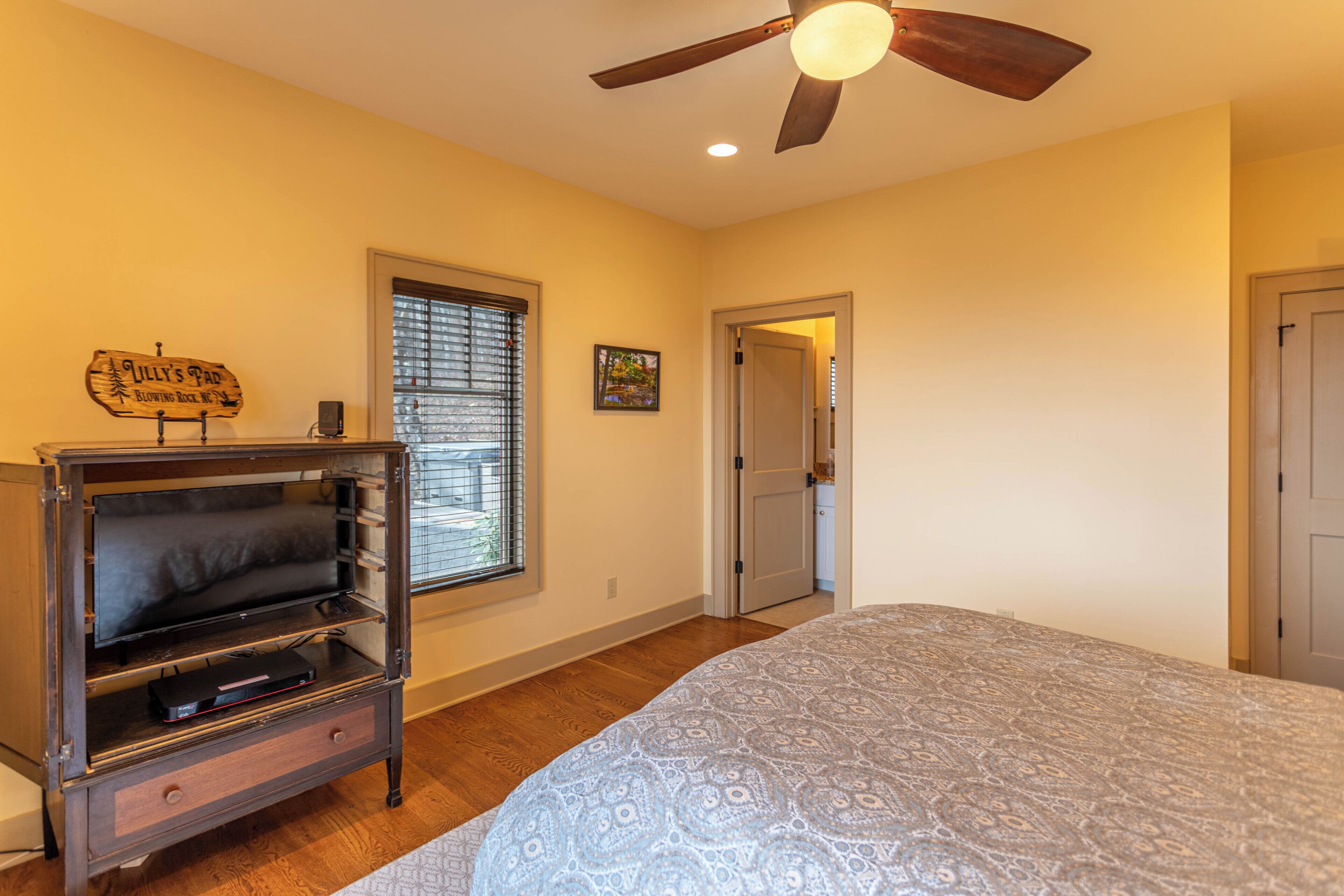 A bedroom with a bed, a wooden cabinet with a television, a ceiling fan, a window with blinds, and a door.