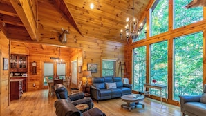 A log cabin interior with a fireplace, leather furniture, and large windows offering a view of the forest.