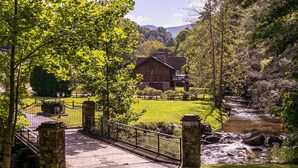 A rustic house with a stone bridge leading to a gated driveway, surrounded by lush greenery and a flowing stream.