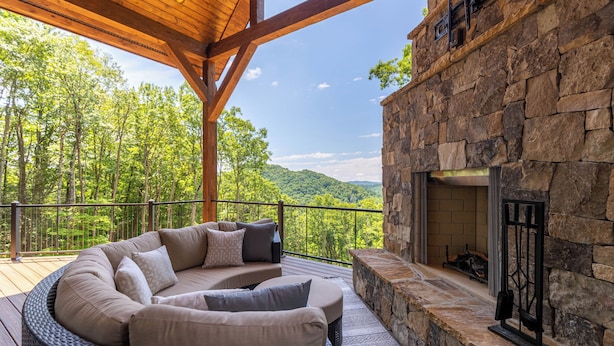 A covered outdoor seating area with a stone fireplace, surrounded by a wooden deck and overlooking a forested landscape. - The Recovery Room at Eagles Nest (Banner Elk)