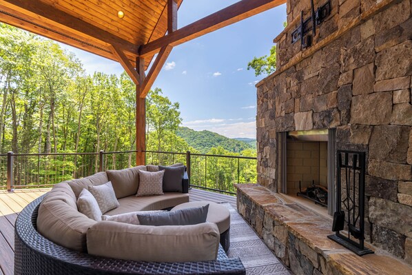 A covered outdoor seating area with a stone fireplace, surrounded by a wooden deck and overlooking a forested landscape. - The Recovery Room at Eagles Nest (Banner Elk)