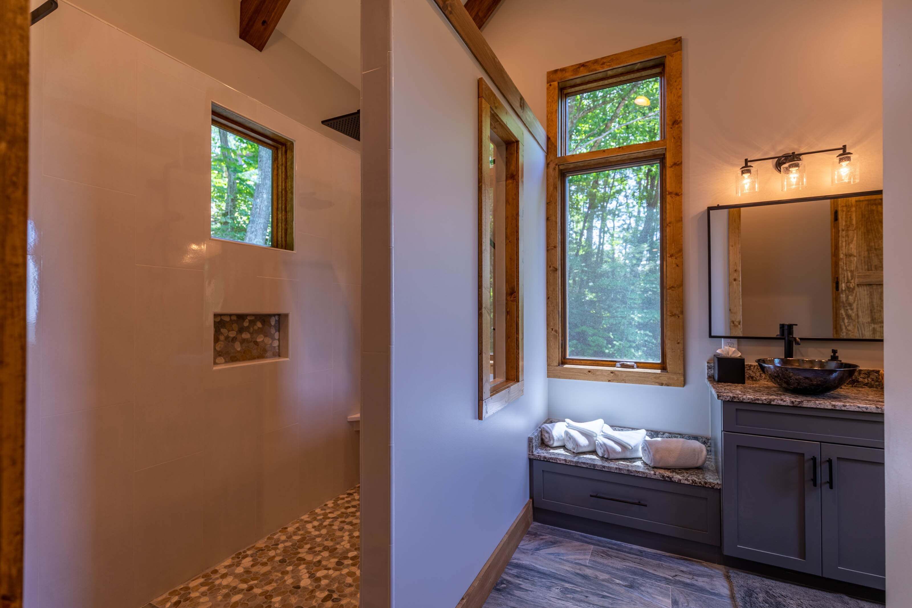 A bathroom with a shower, a sink with a bowl, a mirror, and two windows with views of trees.