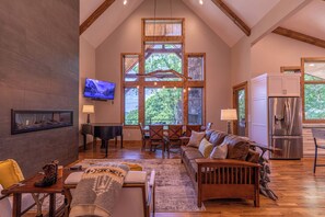 A spacious living room with a high ceiling, wooden beams, a large window with a view of trees, a piano, a sofa, and a dining area. - The Recovery Room at Eagles Nest (Banner Elk)