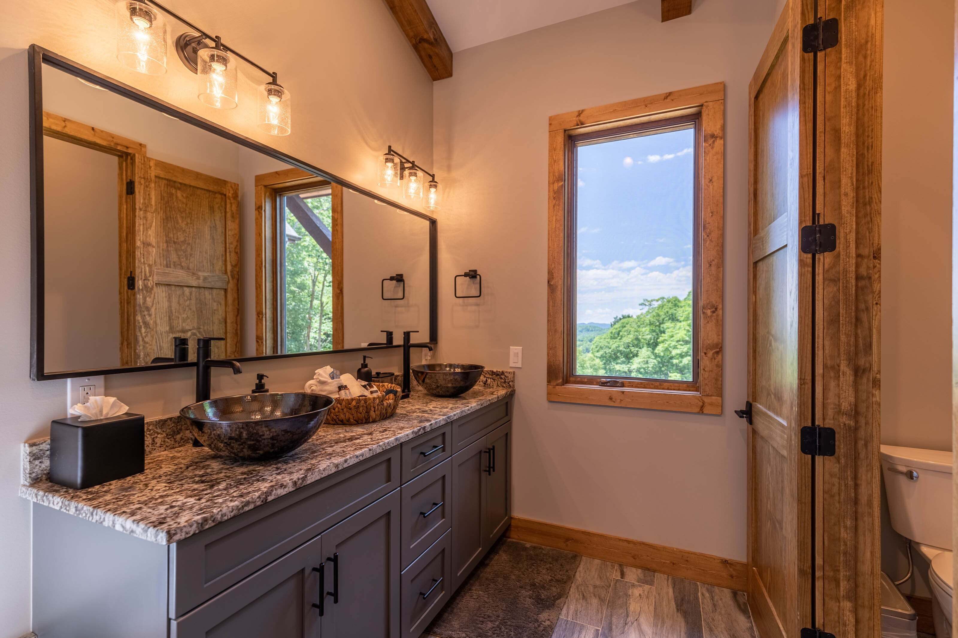 Une salle de bain avec un grand miroir, deux lavabos, une porte en bois et une fenêtre donnant sur un beau paysage.