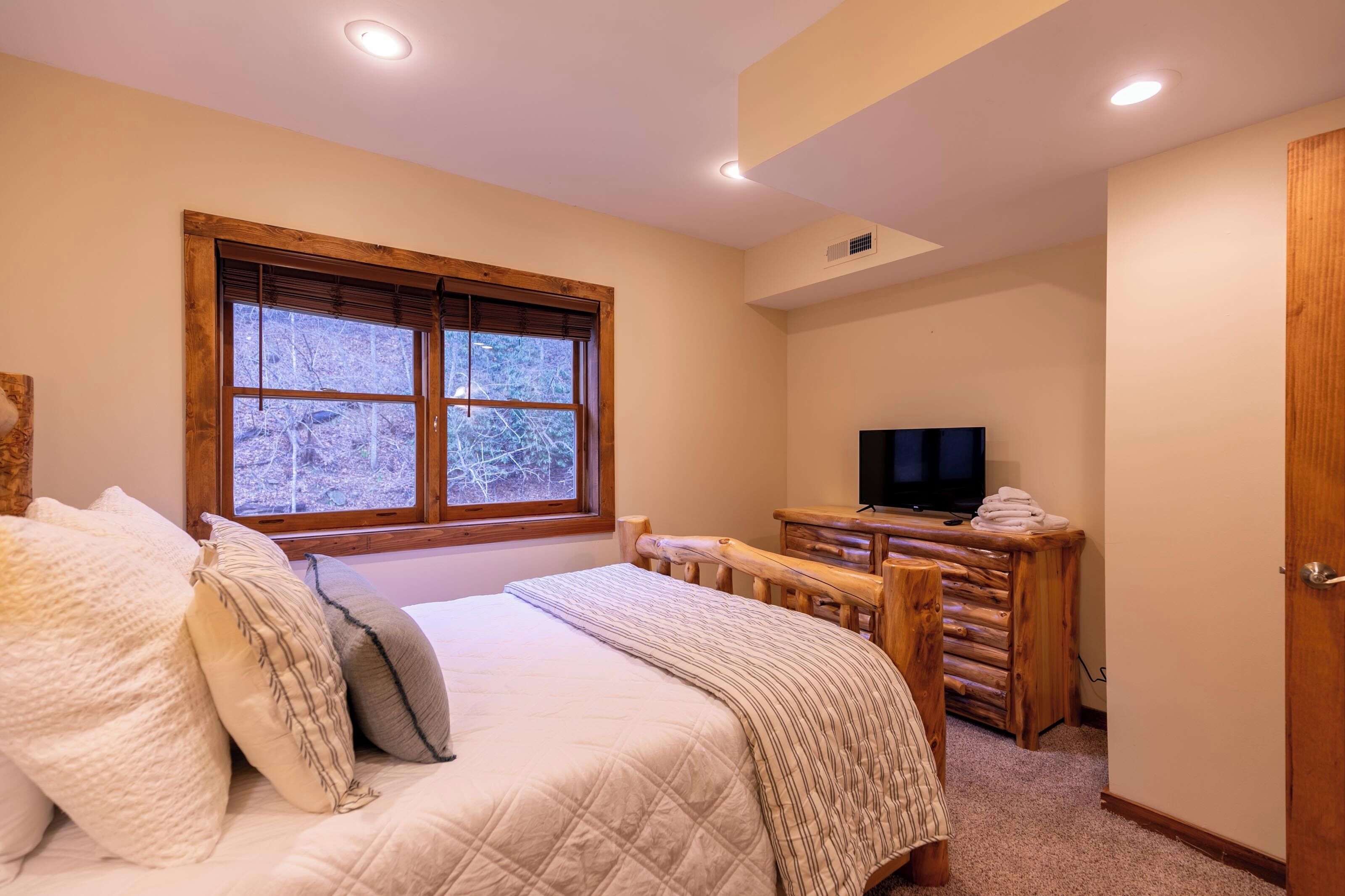 A bedroom with a bed, a television, a wooden bench, and a window with a view of trees.