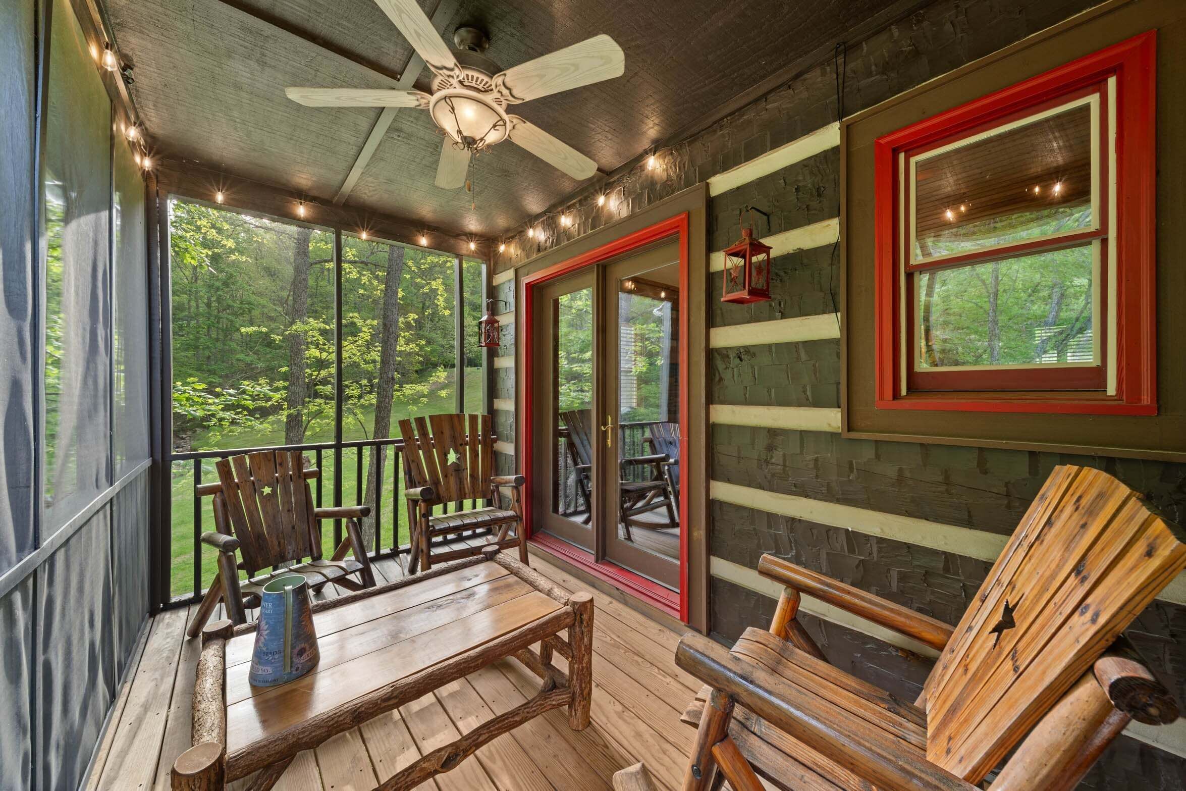 A covered porch with wooden furniture, a ceiling fan, and large windows overlooking a forest.