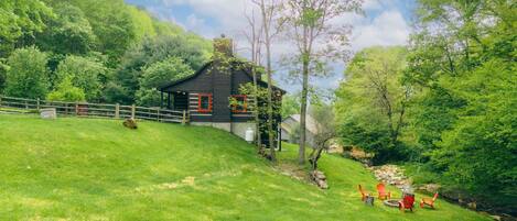 A rustic cabin with a wooden fence, surrounded by lush greenery and a clear sky.