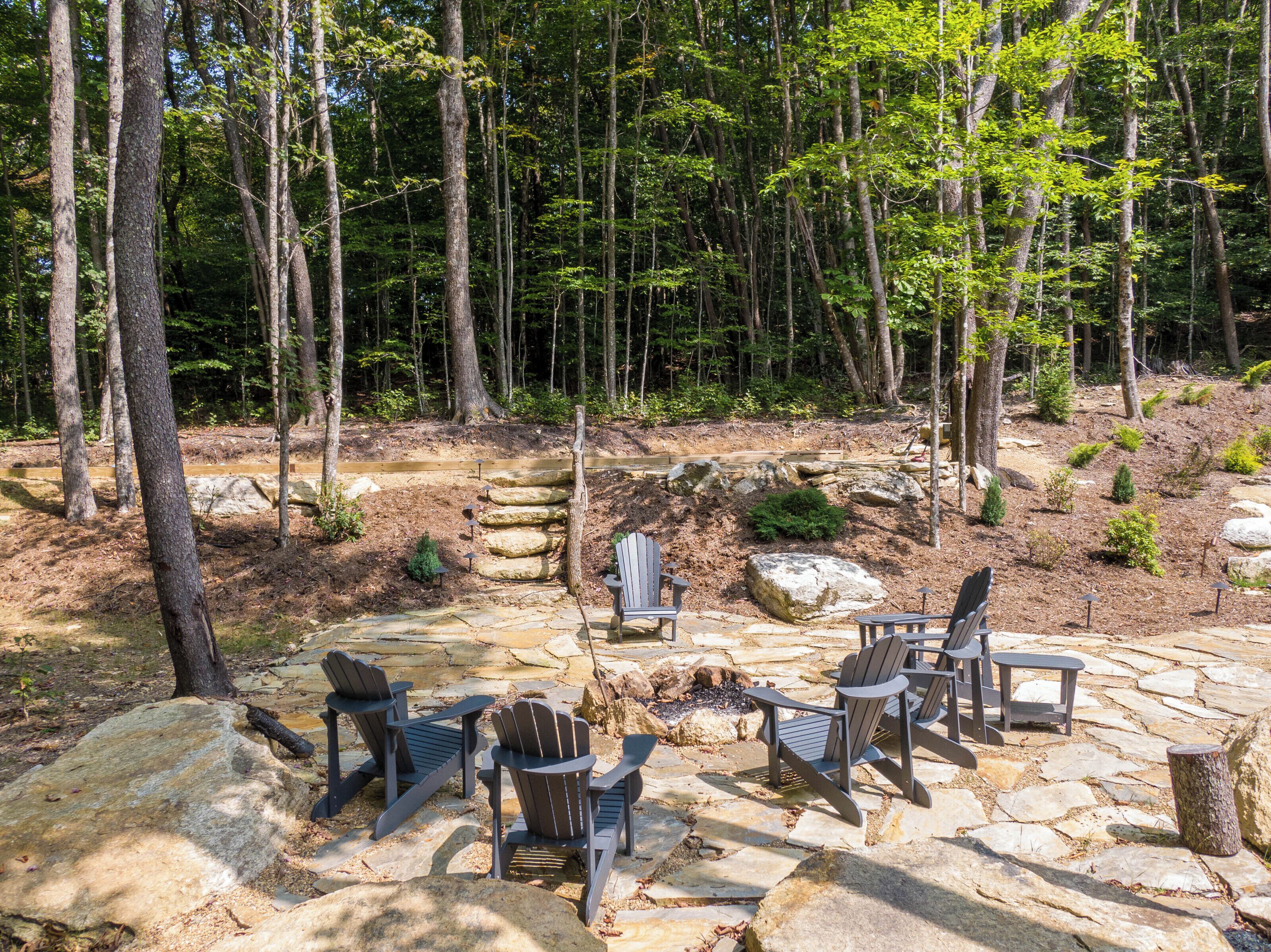 A stone patio with Adirondack chairs and a fire pit in a forested area.