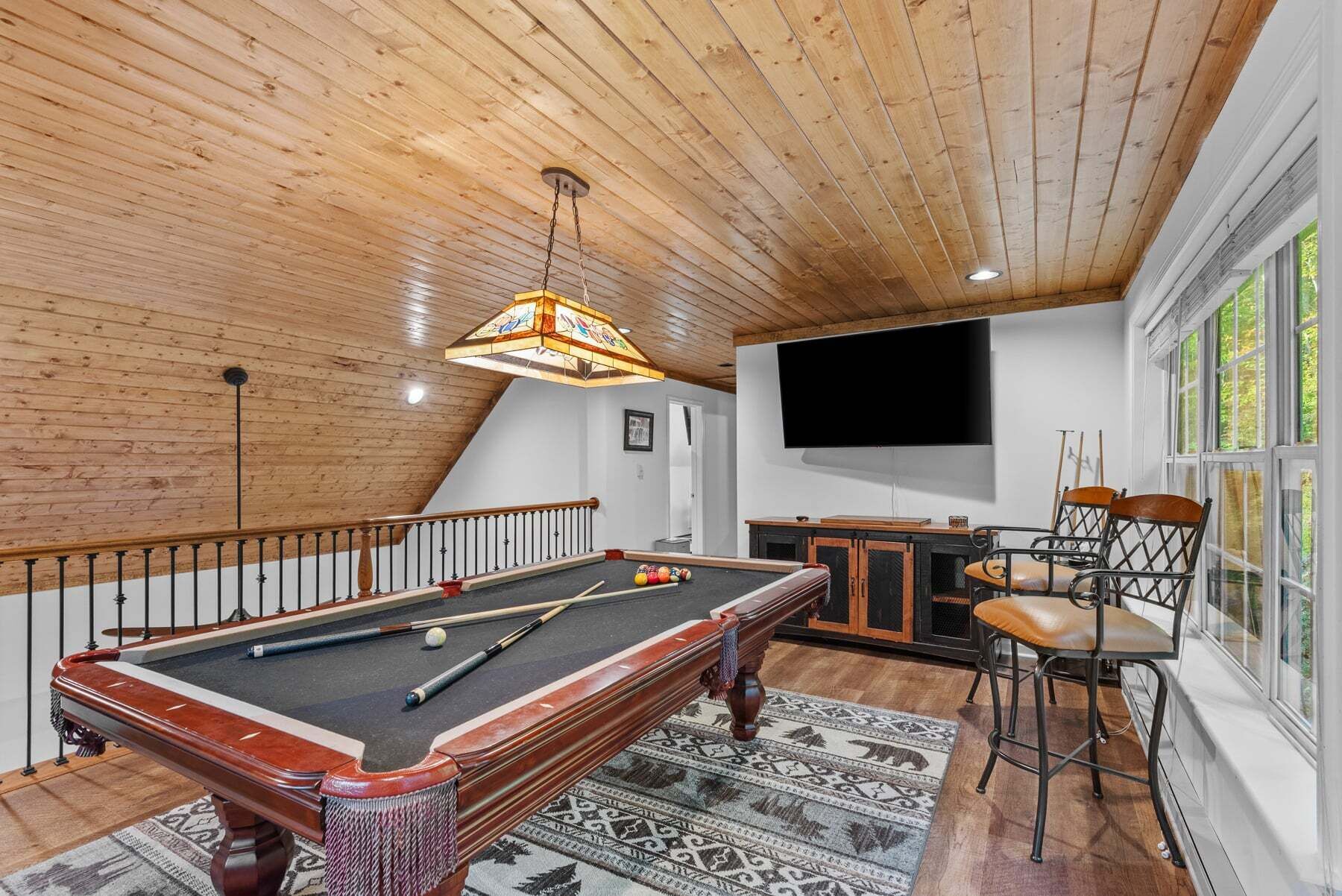 A pool table with balls, a television, and a bar area in a room with wooden ceilings and large windows.