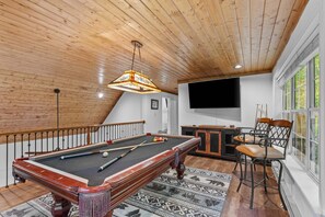 Ein Billardtisch mit Kugeln, ein Fernseher und ein Barbereich in einem Raum mit Holzbalkendecken und großen Fenstern.