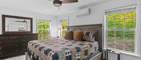 Une chambre à coucher comprenant un lit, une commode, un ventilateur de plafond, de grandes fenêtres et une vue sur la verdure.