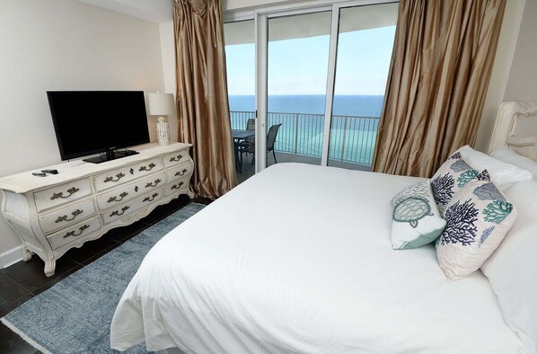 A hotel room with a bed, a TV, a dresser, and a balcony with a view of the ocean.