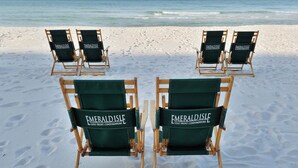 Vier Strandstühle mit dem Schriftzug 'Emerald Isle Gulf Front Condominiums' auf der Rückenlehne, auf einem Sandstrand mit dem Meer im Hintergrund platziert.