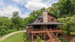 A wooden cabin with a stone chimney, a covered porch, and a staircase leading to the main floor. - Rhododendron Rendezvous (Blowing Rock)