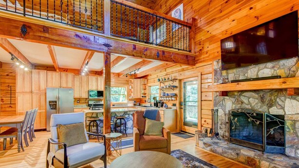 A log cabin interior with a stone fireplace, wooden furniture, a kitchen area, and a balcony. - Rhododendron Rendezvous (Blowing Rock)