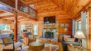 A log cabin interior with a stone fireplace, wooden furniture, a dining area, and a balcony. - Rhododendron Rendezvous (Blowing Rock)