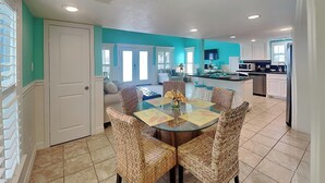 A dining area with a glass table surrounded by wicker chairs, a kitchen in the background, and a teal-colored wall. - Sundancers (Galveston)
