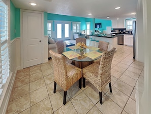 A dining area with a glass table surrounded by wicker chairs, a kitchen in the background, and a teal-colored wall. - Sundancers (Galveston)